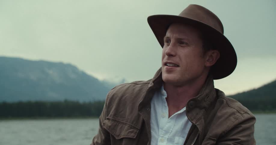 The video shows a man wearing a brown hat and jacket sitting in a boat on a body of water with mountains in the background the mans mouth is moving as if he is speaking and he holds a wooden oar in his hands he looks to his left and smiles then lick