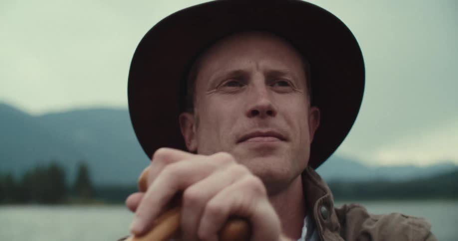 The video shows a man wearing a brown hat and jacket sitting in a boat on a body of water with mountains in the background he is holding a wooden oar the man looks up and to his left as if listening to someone or something he then looks forward again