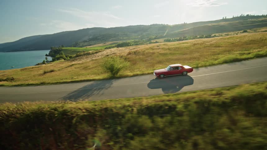 A beautiful vintage red car drives on a road next to a body of water and mountains