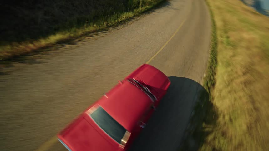 The video shows an aerial view of a red classic ford mustang convertible the car is driving along a winding road that runs next to a large blue lake the surrounding landscape is made up of rolling hills covered in dry golden grass and mountains are vi