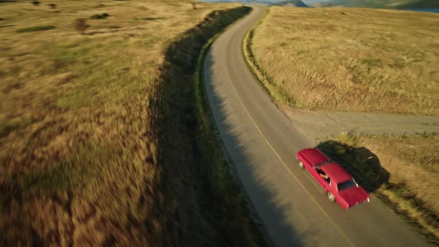 Red car travels through rural fields passing a lake and the mountains
