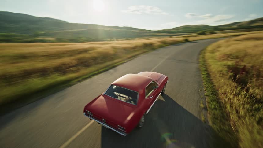 Person drives a red car on a road winding past fields of grass bordering the water and the mountains
