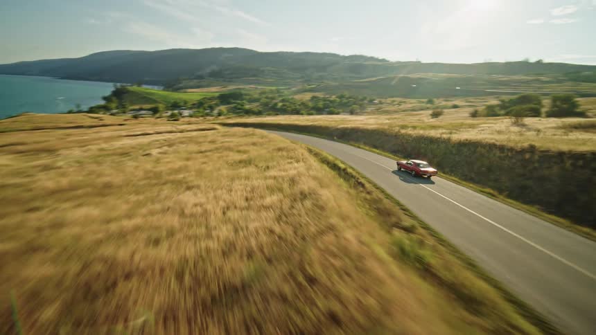 The video shows a bright red classic ford mustang convertible driving down a winding road with an aerial view the road is surrounded by rolling hills covered in golden grass a large blue lake is visible on one side and mountains are in the background