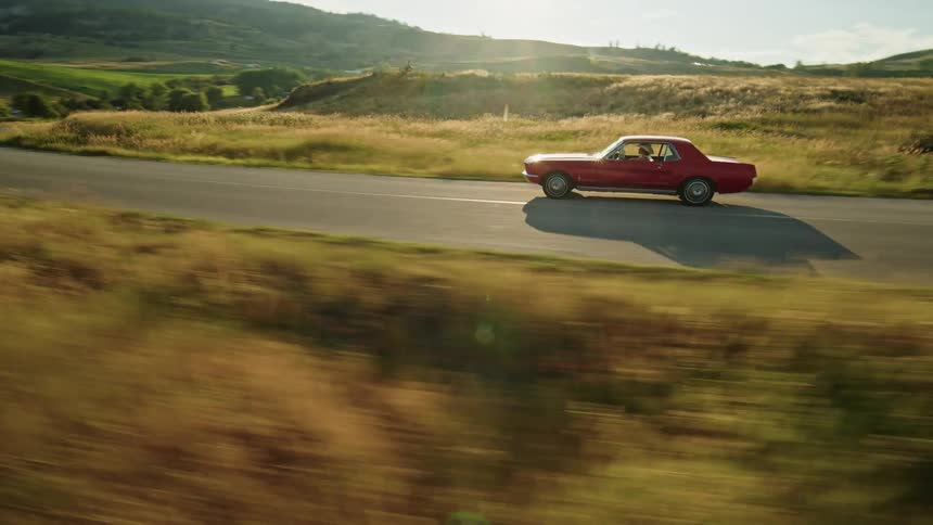 The video shows an aerial view of a red classic ford mustang convertible the camera follows the car as it drives on a winding road next to a large blue lake the road is surrounded by dry golden grass and mountains are visible in the background