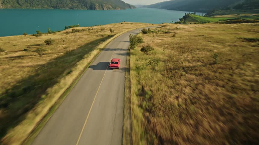 A car drives along a road beside a lake surrounded by grassy hills