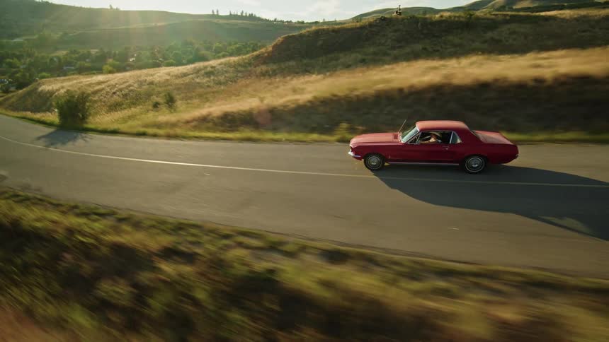 A classic red car drives down a winding road with a single yellow line as it passes by a large body of water and a hillside covered in green and golden grasses under a bright sky