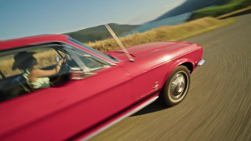 A woman drives a red car along a winding country road near a lake