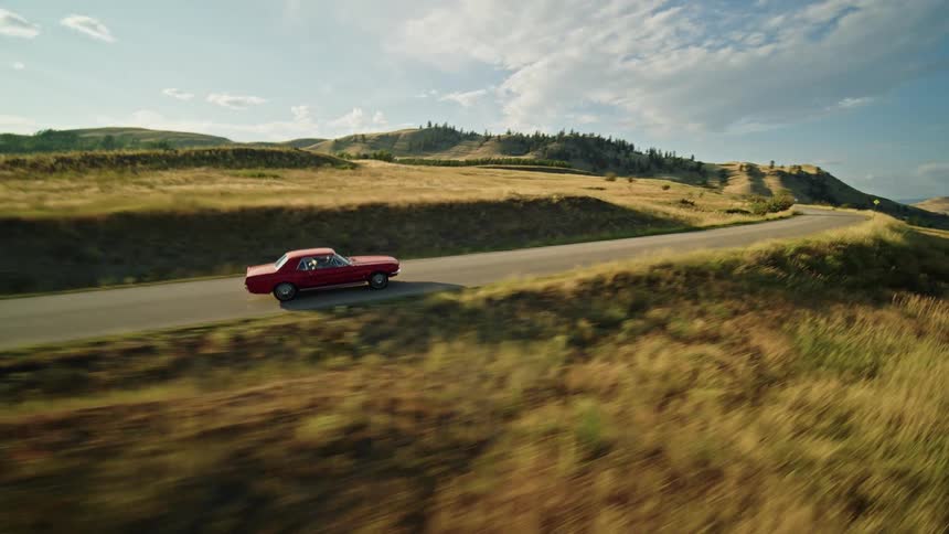 A red car driving on a lonely road in a rural area flanked by grasslands on both sides at midday
