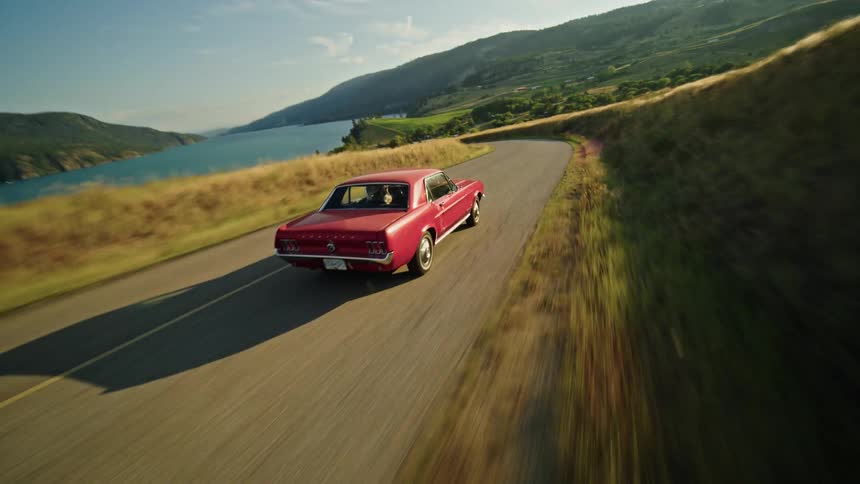 The video shows an aerial view of a red classic ford mustang convertible driving down a winding road the car is surrounded by rolling hills covered in golden grass with a large blue lake and mountains in the background the sun is low in the sky
