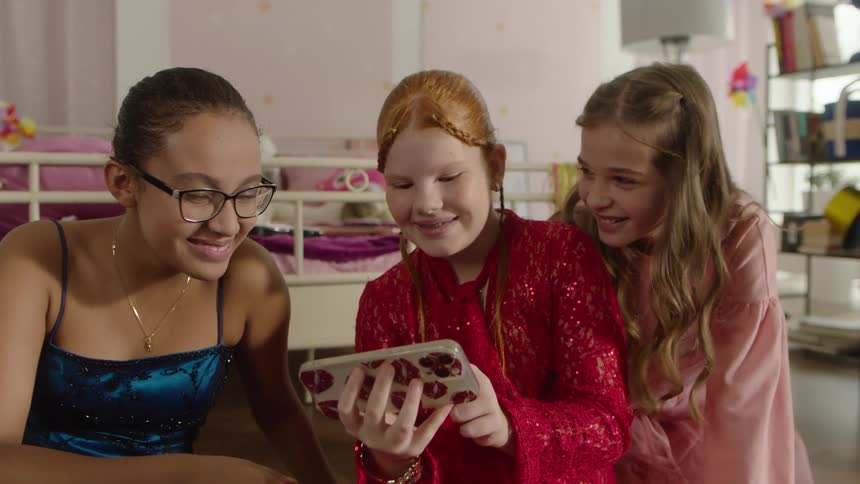 Three girls in party outfits laugh while looking at a phone