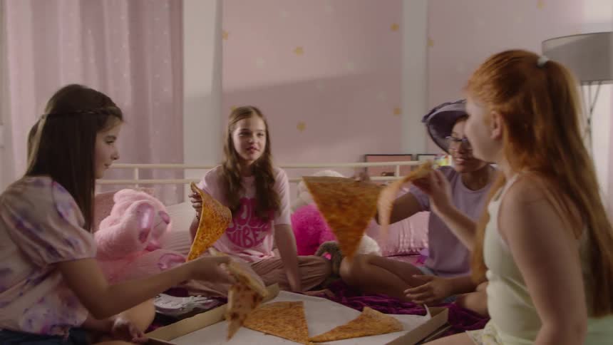 Friends sharing a pizza while sitting on a bed toast each other with large slices