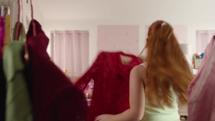 Three teenage girls one picking a piece of clothing from a hanger and showing it to her two friends sitting on the bed as they cheer her on