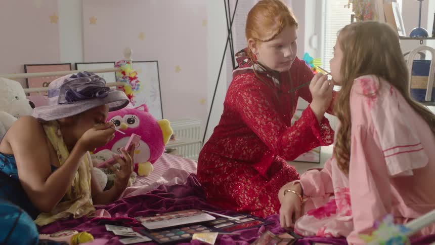 Girls wearing fancy dresses apply makeup while in a bedroom during the daytime