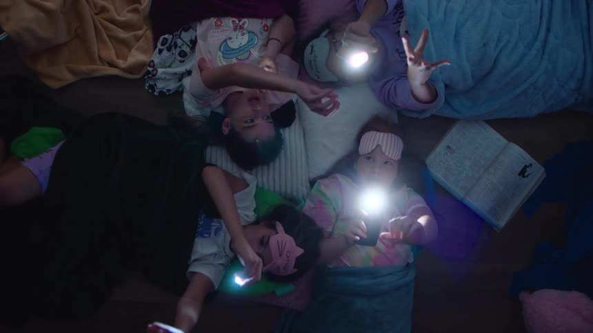 Four girls lying on a floor and playing with flashlights all with pajamas wearing sleeping masks and blankets