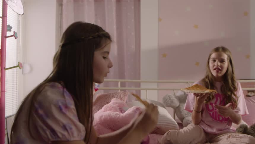 Young female friends sitting on a bed together during the daytime eat pizza