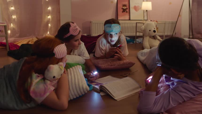 A group of teenage girls are on the floor with sleep masks on their heads and flashlights under their chins