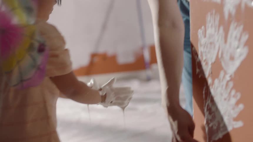 A little girl wearing a dress with fairy wings dips her hands into a tray of white paint she then presses her paintcovered hands against a wall leaving a trail of messy white handprints