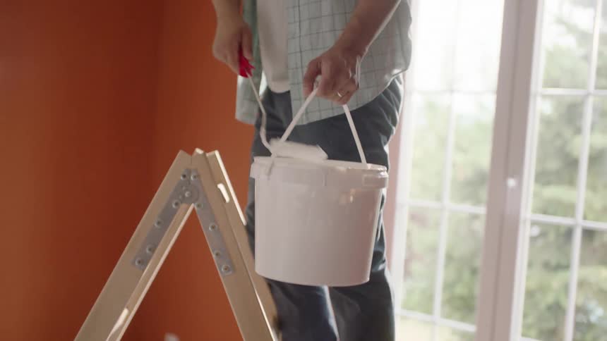 A man paints the ceiling with a roller while standing on a ladder