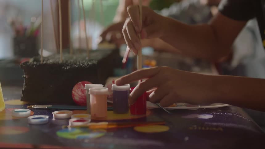 A boy sits at a table and paints a small sphere on a stick while a person sits behind him