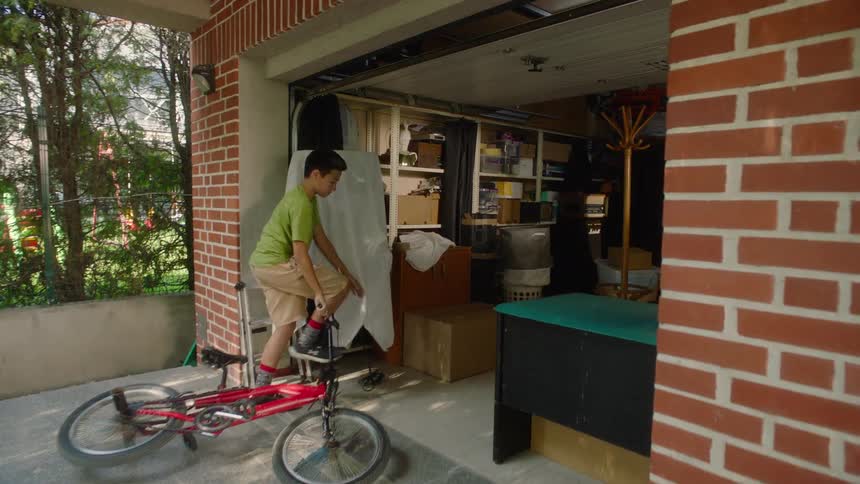A mother carrying a child moves a box from the garage into the back of a truck while a teenage boy arrives on a bike and heads to the garage to pick up a box