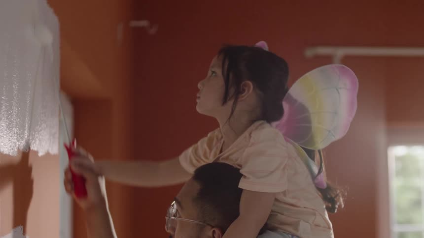 Daughter sits on the shoulders of her father while they paint the wall