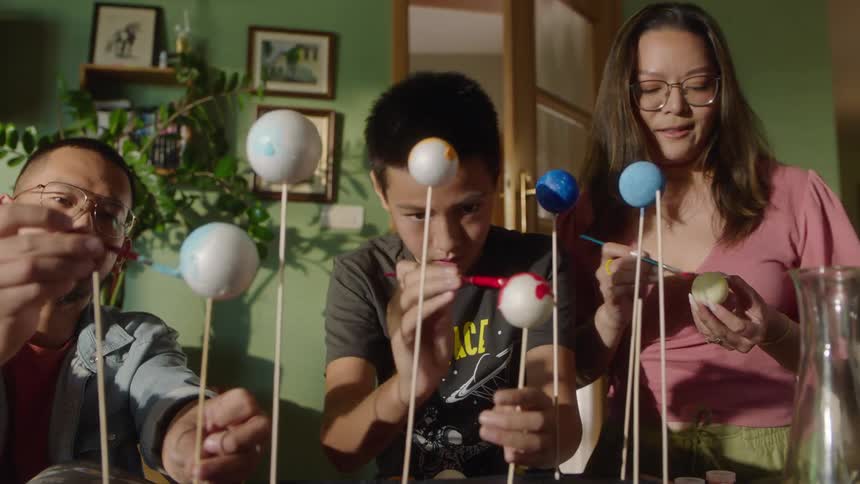 A man a woman and a boy sit at a table and paint spheres on sticks