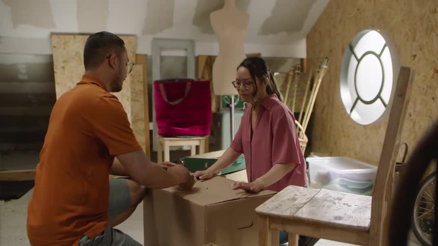 The video shows three people in what appears to be an attic or workshop space with shelves and boxes visible in the background one woman is seen placing a bowl on a shelf while two men are busy packing up boxes on the floor