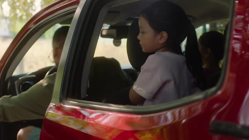 A man opens a door and gets into a red car where a child and a woman sits