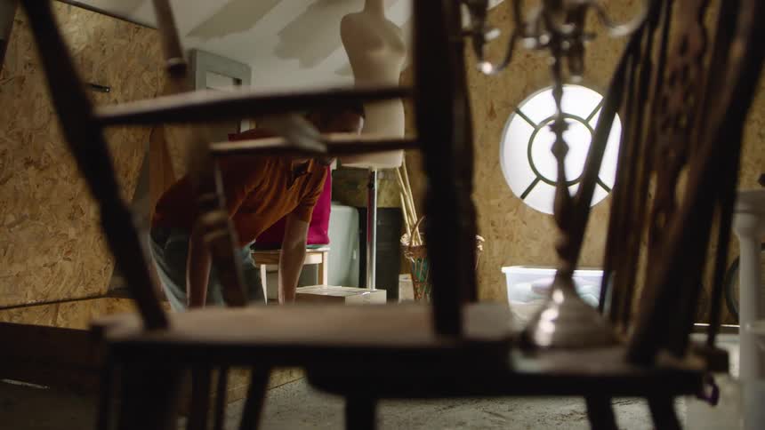 A man wearing a rust colored shirt and shorts crouches and moves through a cluttered attic filled with old furniture and a mannequin