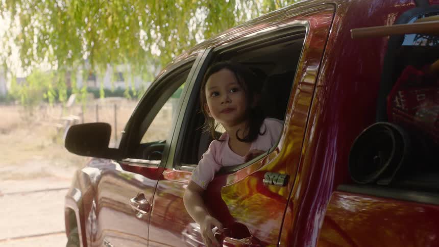 A father dressed in a green shirt and brown pants opens the door to a red truck and looks into the back seat where his young daughter is sitting and looking out the window