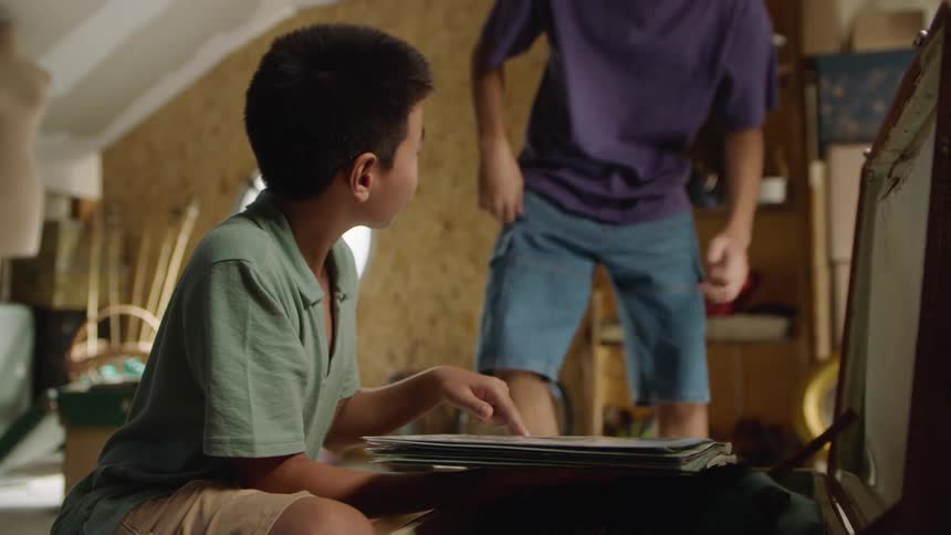Two boys in an attic space open a trunk and look through a book inside it
