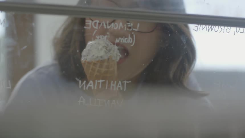 A woman eats an ice cream cone while looking at the flavors
