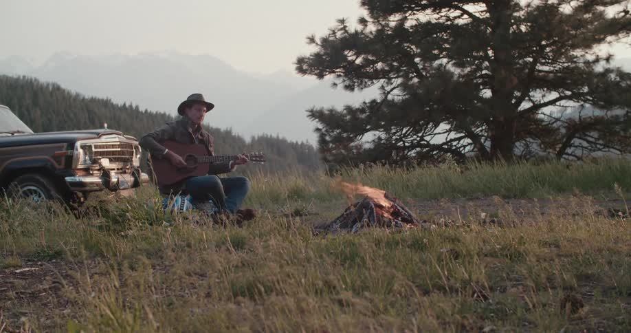 A middleaged man wearing a hat sits on a grassland by a campfire playing a guitar with his car parked behind him at sunset