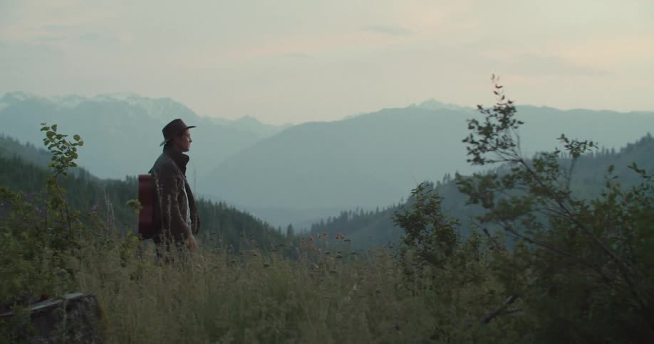 The video shows a man with a hat and a backpack standing on a grassy hill looking out at a mountain range the mountains create two distinct horizontal lines a lower line of forested hills and a higher fainter line of snowcapped peaks in the distance