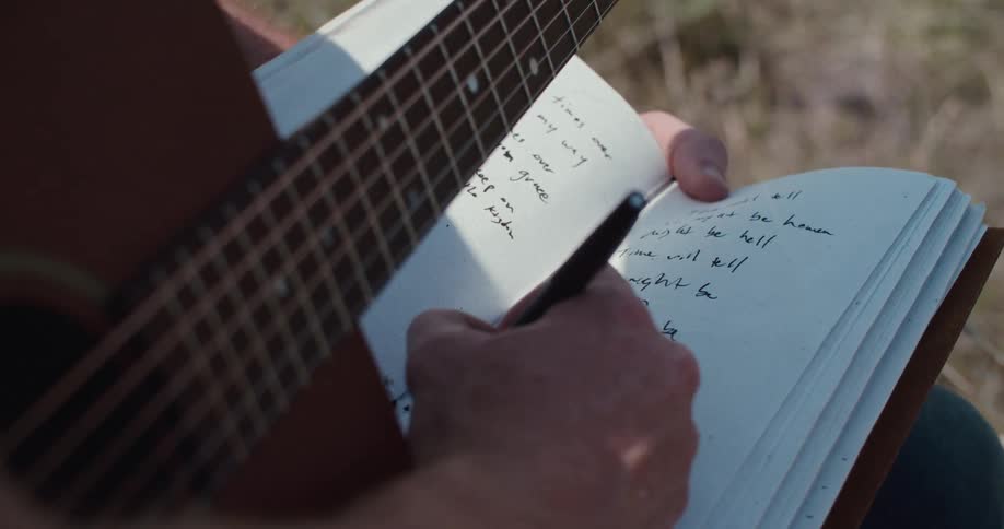 Person writing in a notebook while sitting holding a guitar