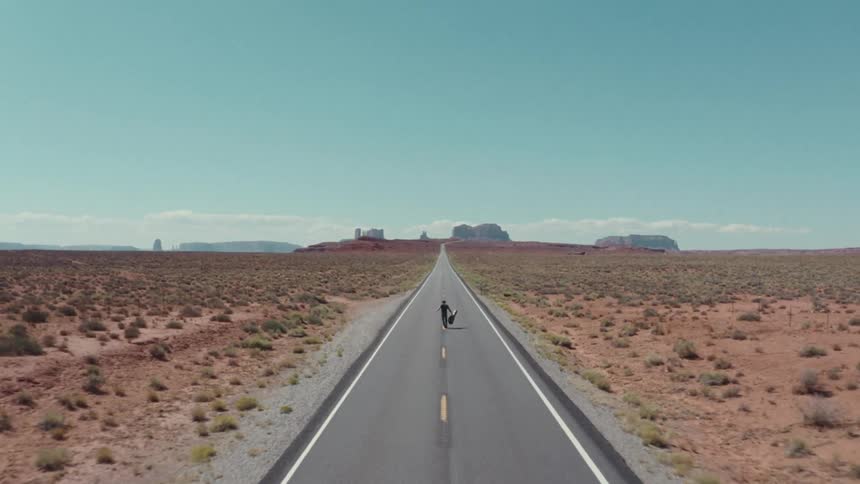 A person walks freely down a long desert highway carrying a guitar