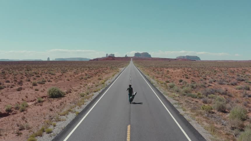 A man with a guitar case walks down the center of an empty desert highway the long straight road is surrounded by dry brush and distant rock formations under a clear bright blue sky