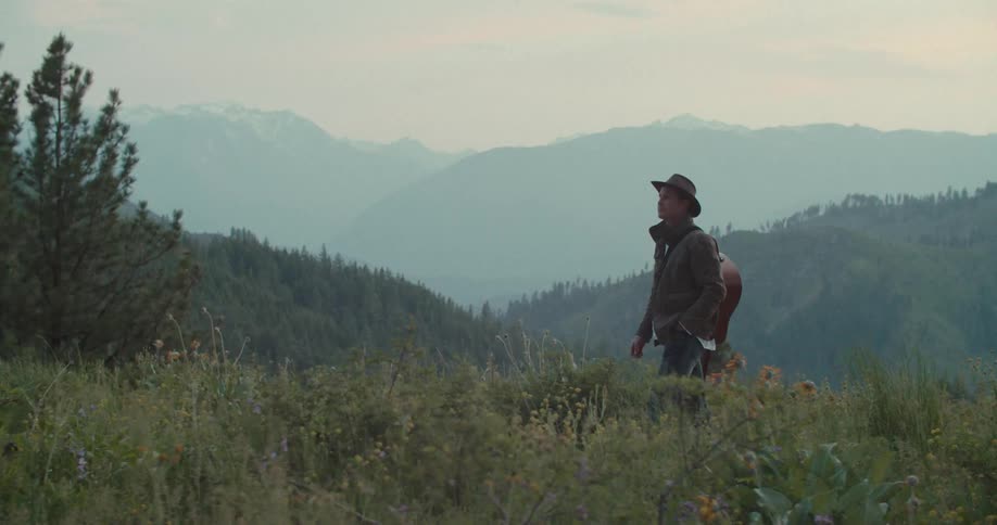 A man walks on a grassy mountain with a backpack overlooking distant peaks