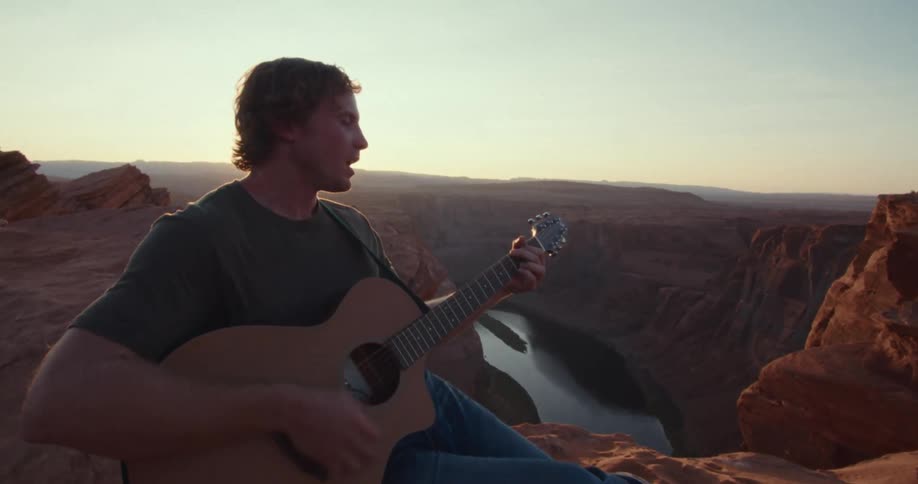 A man sits on the edge of a redrock canyon playing an acoustic guitar as the sun sets he is looking out over a river far below and the low sunlight casts a warm glow on his face