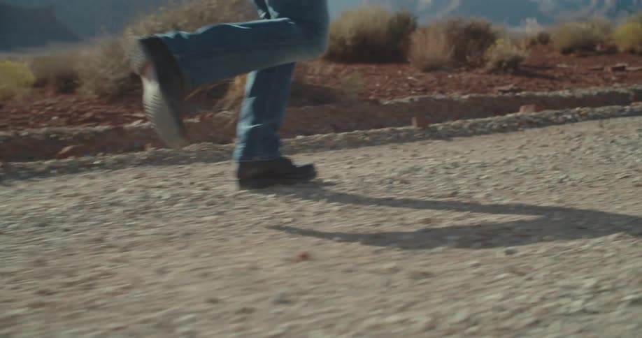 A person in jeans and black shoes runs along a dusty path casting long shadows
