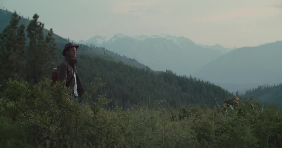 A man carrying a guitar walks through the mountain landscape at dusk