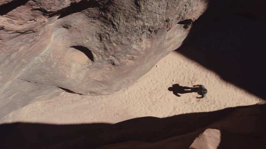 An overhead shot looks down on a person walking along a sandy canyon floor the sun casts a stark shadow and the persons figure is dwarfed by the massive rock formations