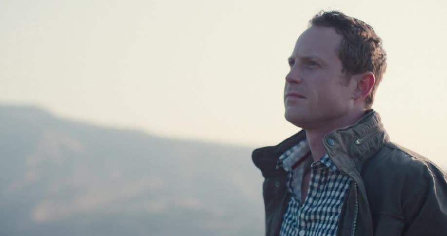 A closeup side profile shot of a man wearing a dark hat a green jacket and a plaid shirt he looks out over a foggy landscape then takes his hat off and puts it back on