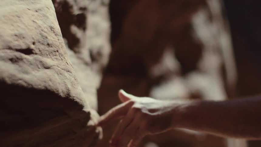 A persons bare hand reaches out and runs their fingers along the rough textured surface of a sunlit rock wall inside a cave or canyon