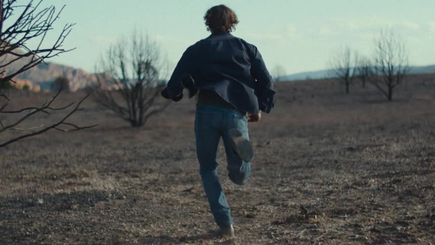 A man wearing jeans boots and a black shirt running on sand in the wild at midday