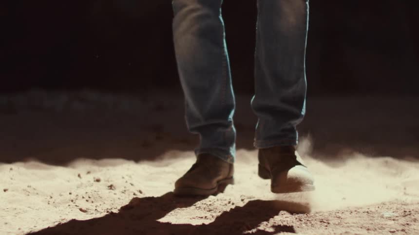 Two feet in worn brown boots and faded blue jeans walk across a sandcovered ground as the person walks the sand kicks up in small puffs of dust with each step