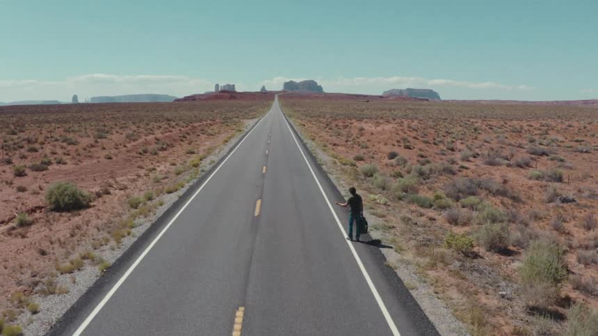 A man stands on a desert highway hitchhiking for a ride