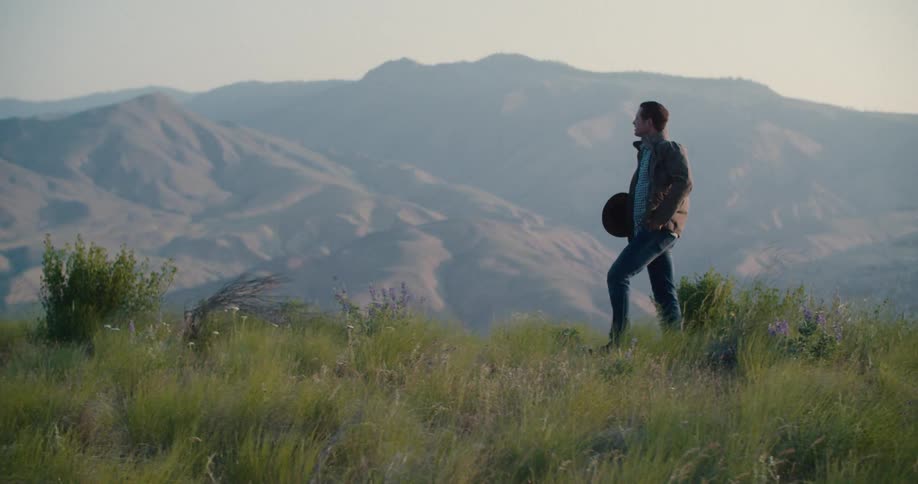 A man in a hat and a jacket walks on a grassy hilltop briefly holding his hat with one hand and then removing it with both hands as he continues walking across the field with a mountain range in the distance