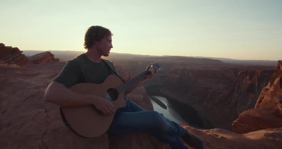 A man sits on the edge of a tall cliff and plays a guitar while looking out over a deep canyon and a winding river as the sun sets behind him