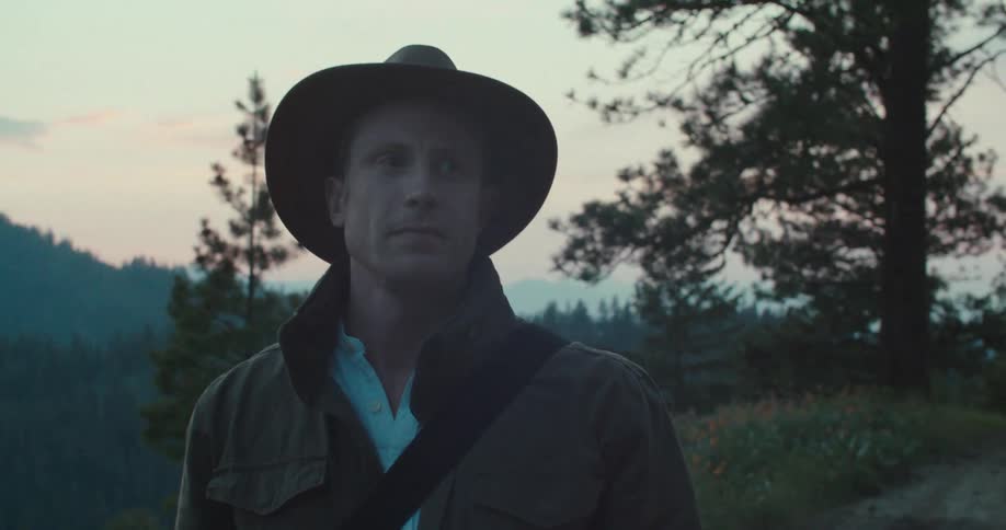 A side profile of a man in a hat looks out at a mountainous landscape he then turns his head to look directly at the camera with a serious and contemplative expression